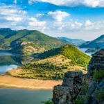 Person stands on a rocky cliff overlooking a winding lake and green mountains under a partly cloudy sky.