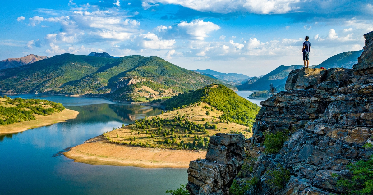 Person stands on a rocky cliff overlooking a winding lake and green mountains under a partly cloudy sky.