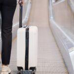 Person in black pants pulling a white suitcase on an airport moving walkway
