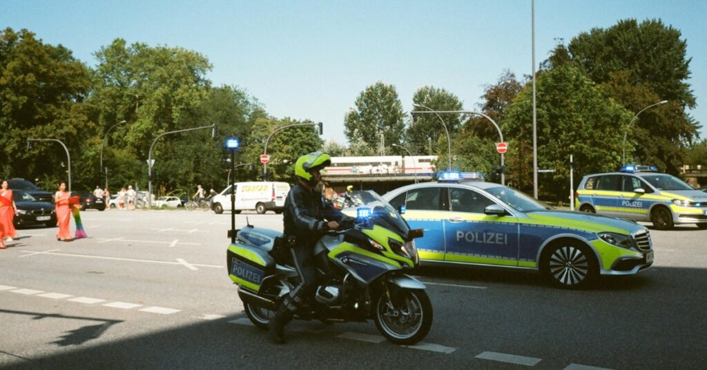 Police motorcycle and patrol car with flashing lights block a city intersection as pedestrians cross nearby