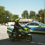 Police motorcycle and patrol car with flashing lights block a city intersection as pedestrians cross nearby