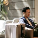 A business traveller sits in a hotel lounge reading beside a large suitcase.
