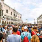 Crowds of tourists fill Venice’s Piazza San Marco between ornate historic buildings under a cloudy sky