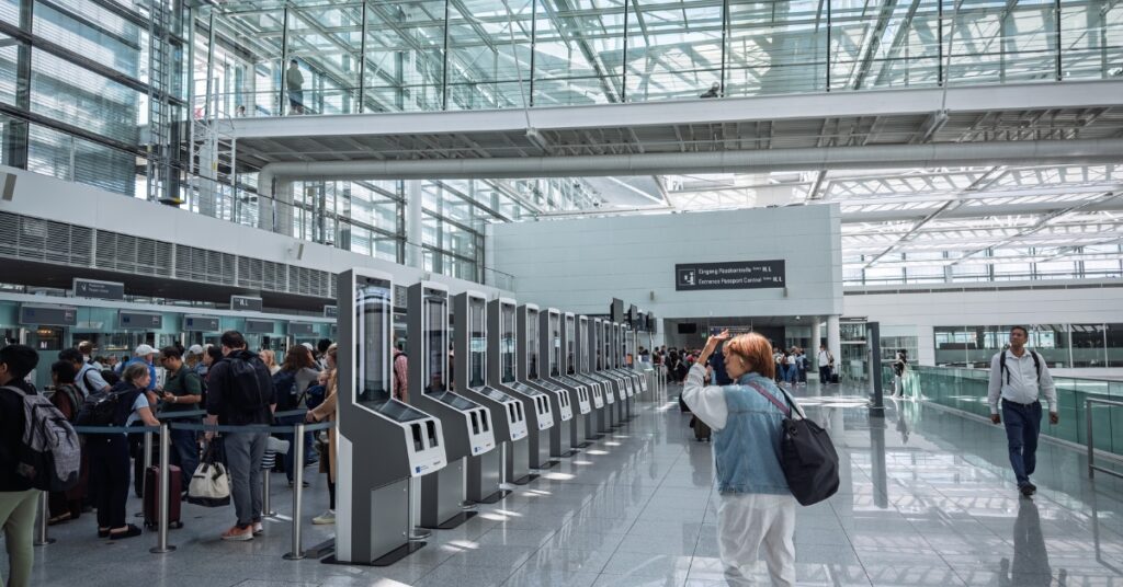 Travellers queue at self-service passport control kiosks inside a bright airport terminal with a glass walkway overhead