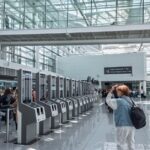 Travellers queue at self-service passport control kiosks inside a bright airport terminal with a glass walkway overhead