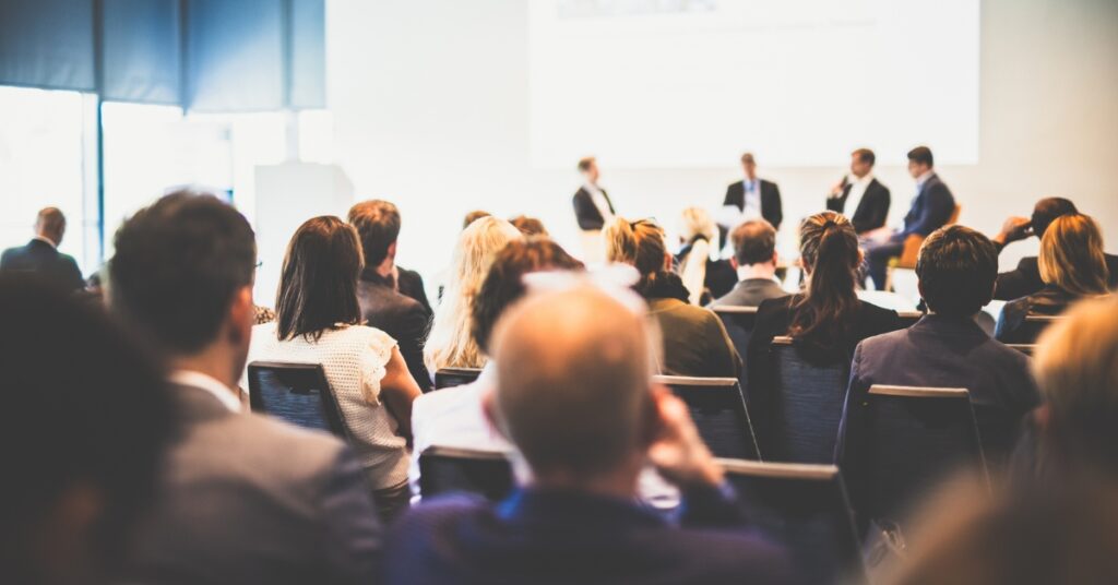 Audience attending a business conference with panellists speaking on stage in a bright conference room