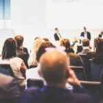 Audience attending a business conference with panellists speaking on stage in a bright conference room
