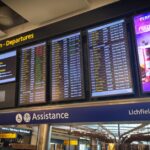 Airport departure information boards display flight schedules and gate details above a terminal concourse