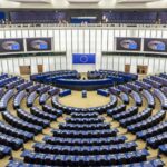 European Parliament plenary chamber with curved rows of seats and EU flags displayed above the central podium