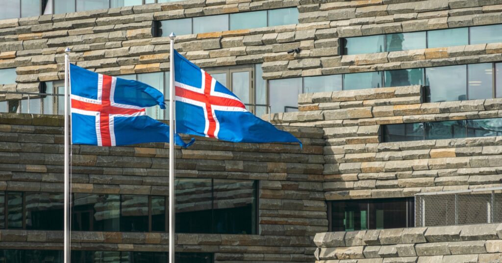 Two Icelandic flags fly outside a modern stone-and-glass building