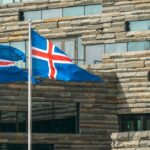 Two Icelandic flags fly outside a modern stone-and-glass building