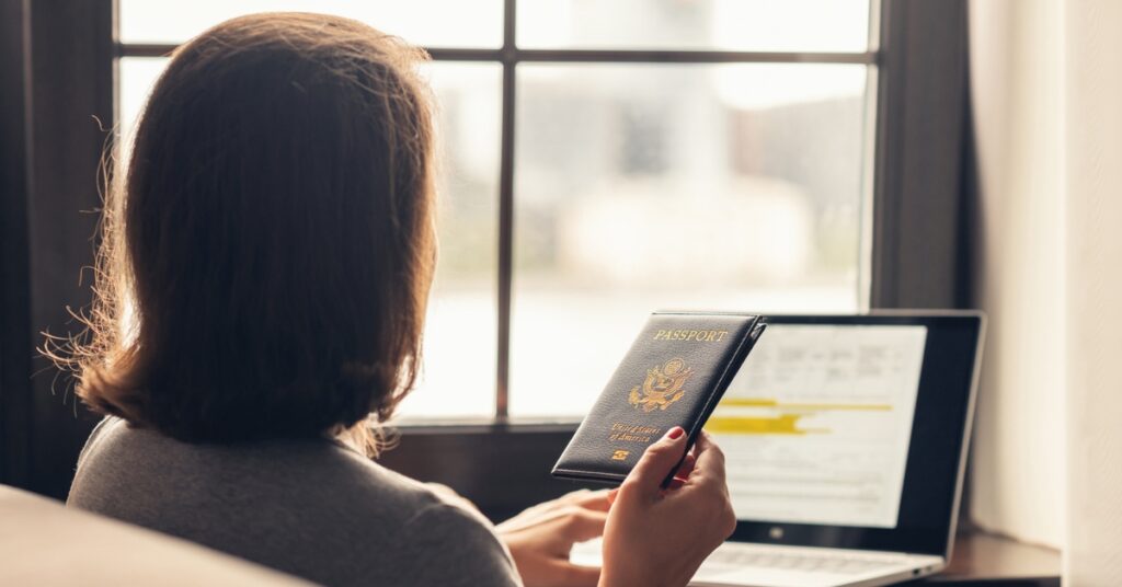 Person holds a passport while using a laptop by a window, preparing travel documents online