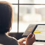 Person holds a passport while using a laptop by a window, preparing travel documents online