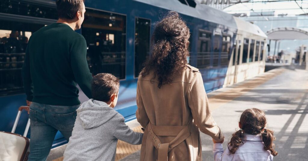 Two adults and two children walk along a station platform beside a blue train with luggage