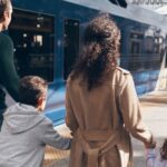 Two adults and two children walk along a station platform beside a blue train with luggage