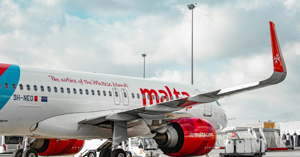 Air Malta aircraft parked on the airport tarmac beneath a cloudy sky, with ground service equipment nearby