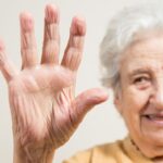 Smiling elderly woman raises an open hand towards the camera