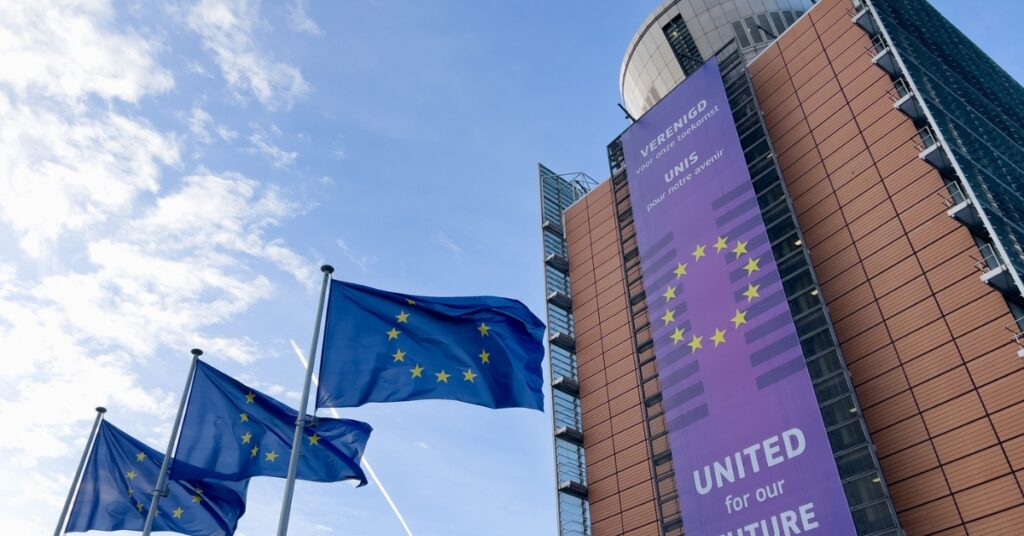 European Union flags fly outside a modern building displaying a purple ‘United for our Future’ banner