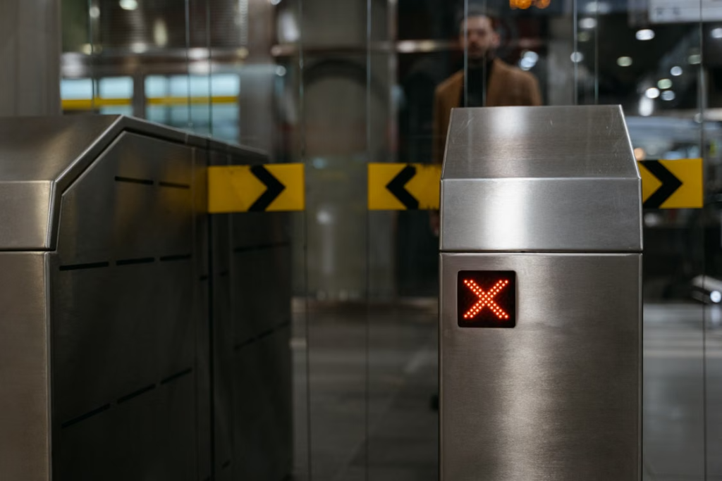 Metal ticket barrier with a red X indicator at a station entrance, with a person visible behind the glass