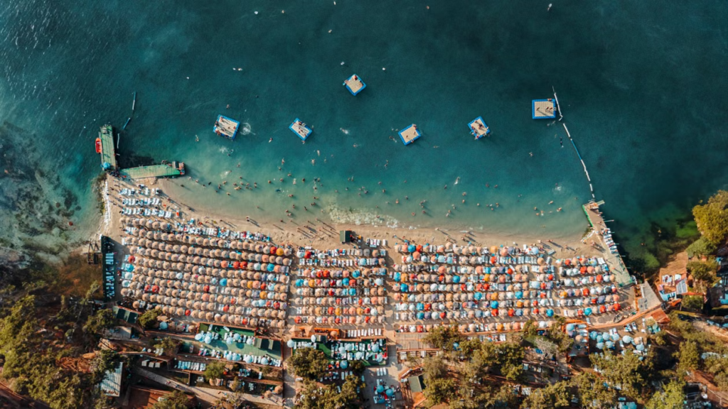 Aerial view of a crowded beach lined with colourful parasols, with swimmers and floating platforms offshore