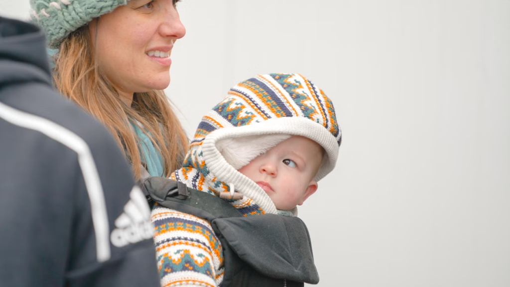 A baby in a patterned hooded jumper is carried in a front carrier beside a smiling woman