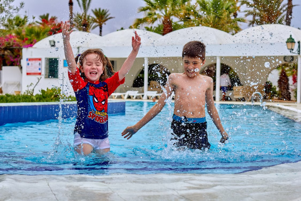 Two children splash and play in a swimming pool surrounded by palm trees