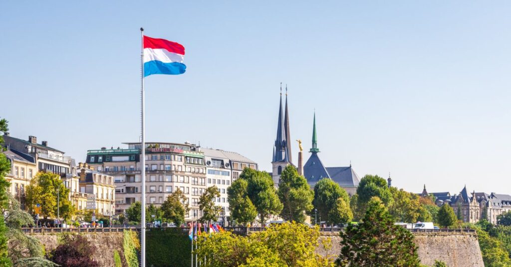 Luxembourg flag flying above gardens and city buildings, with church spires rising against a clear blue sky