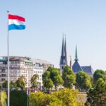 Luxembourg flag flying above gardens and city buildings, with church spires rising against a clear blue sky