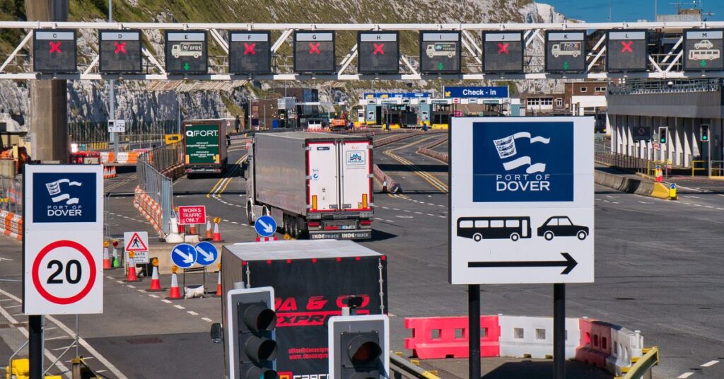 Vehicles queue at Port of Dover check-in lanes beneath overhead signs, with white cliffs in the background