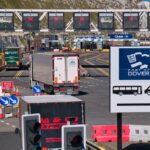 Vehicles queue at Port of Dover check-in lanes beneath overhead signs, with white cliffs in the background