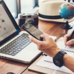 Person plans a trip using a laptop and phone beside a notebook, globe, camera and model aeroplane