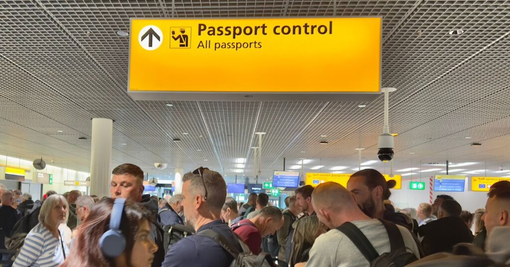 Crowd of travellers queue beneath a yellow Passport control sign for all passports at an airport