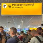 Crowd of travellers queue beneath a yellow Passport control sign for all passports at an airport