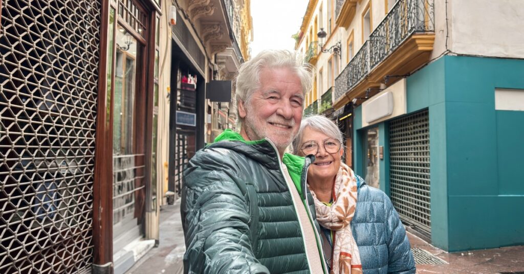 Older couple smiling for a selfie on a narrow European street lined with colourful buildings and balconies