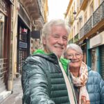 Older couple smiling for a selfie on a narrow European street lined with colourful buildings and balconies