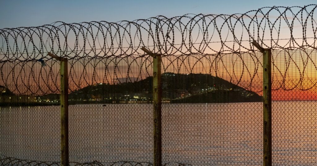 Razor wire lines a high border fence beside the water at sunset, with a lit hillside in the distance