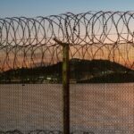 Razor wire lines a high border fence beside the water at sunset, with a lit hillside in the distance
