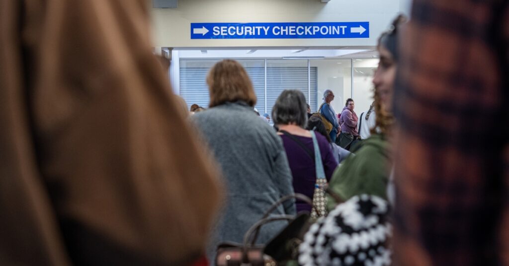 Travellers queue beneath a security checkpoint sign at an airport terminal