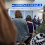 Travellers queue beneath a security checkpoint sign at an airport terminal