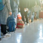 Travellers queue with suitcases and backpacks inside a bright airport terminal