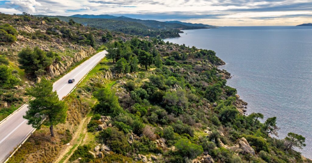 Winding coastal road through green rocky hills beside a calm blue sea under a cloudy sky