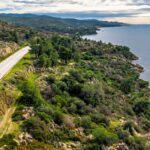 Winding coastal road through green rocky hills beside a calm blue sea under a cloudy sky