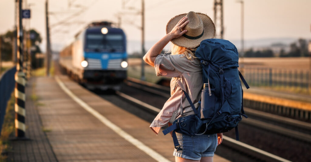A woman wearing a straw hat and carrying a large blue backpack watches an approaching train at a rural railway station