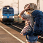 A woman wearing a straw hat and carrying a large blue backpack watches an approaching train at a rural railway station
