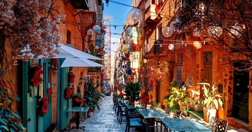 Colourful café tables line a narrow stone street between historic buildings, with plants and hanging decorations overhead