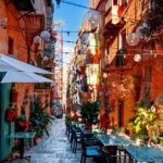 Colourful café tables line a narrow stone street between historic buildings, with plants and hanging decorations overhead
