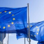 European Union flags waving on flagpoles outside modern buildings under a clear blue sky