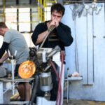 Two glassblowers shape molten glass using specialised equipment inside an industrial glassmaking workshop