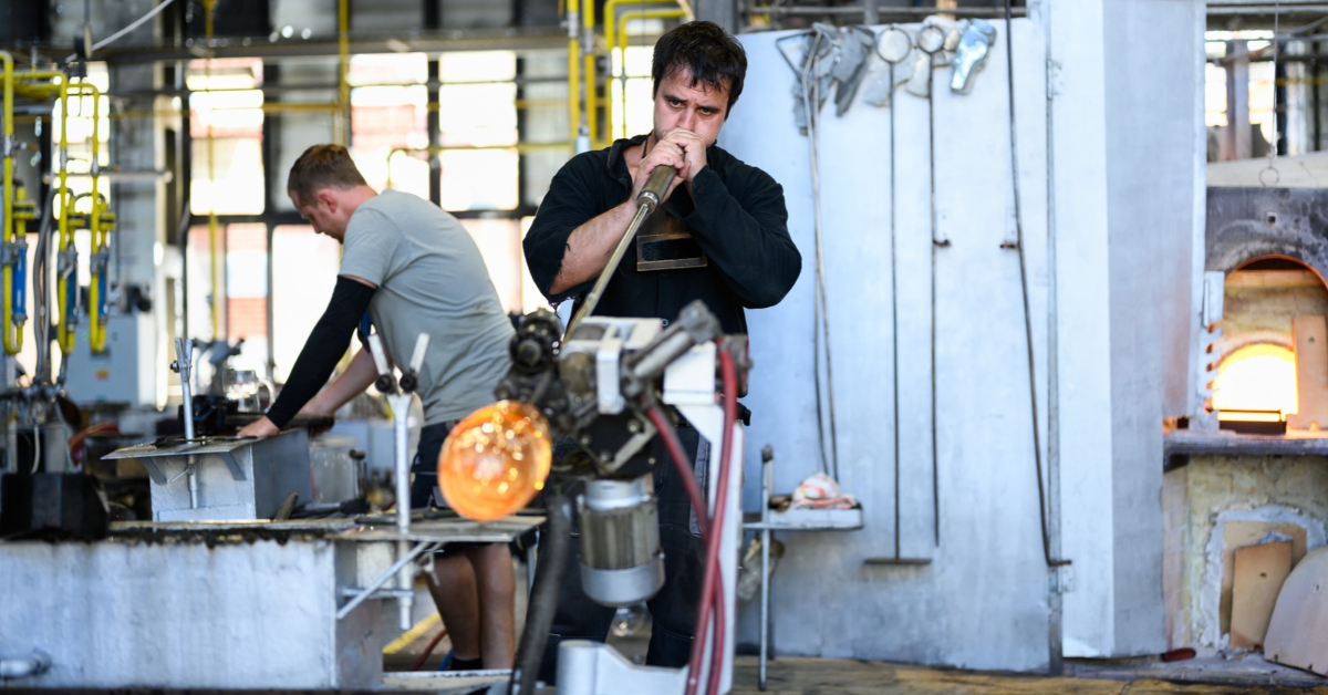 Two glassblowers shape molten glass using specialised equipment inside an industrial glassmaking workshop