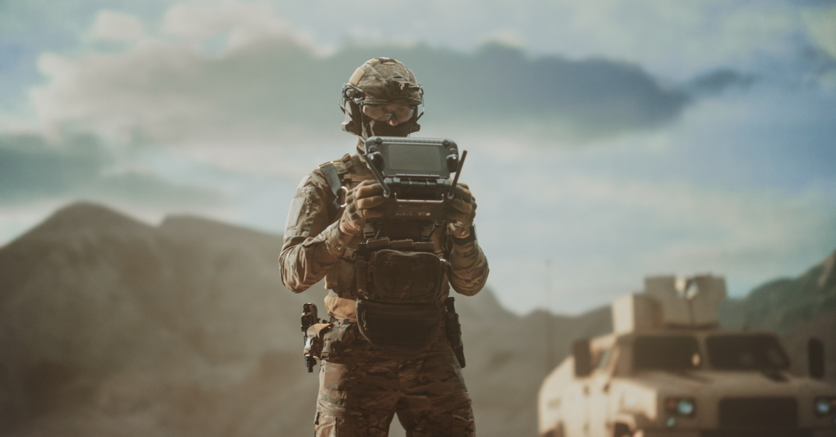 Soldier in camouflage uniform operating a drone controller, with an armoured military vehicle and mountains in the background